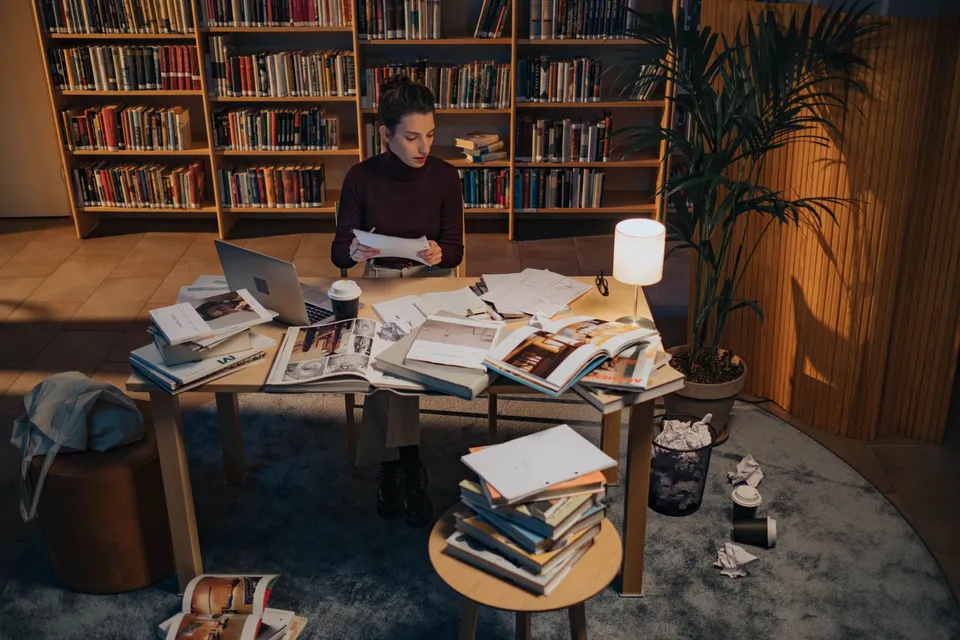 An adult working late at a cluttered table in a library, surrounded by books and paperwork.